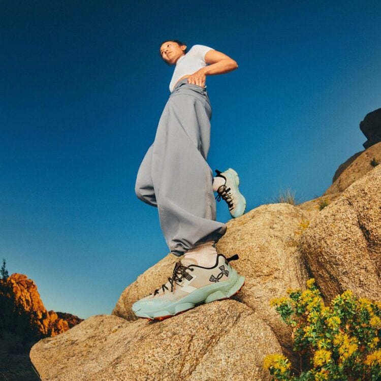 Close-up of an orange trail running shoe on a rocky ledge with a blue sky in the background, person wearing black socks and shorts partially visible beneath the rock.