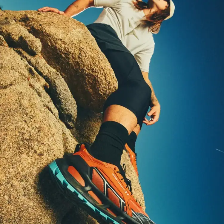 Close-up of an orange trail running shoe on a rocky ledge with a blue sky in the background, person wearing black socks and shorts partially visible beneath the rock.